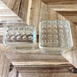 May include: Two rectangular clear glass storage containers with lids. Each lid features a grid of raised circular bumps on the top surface. The smaller container is on the left, and the larger container is on the right, both made of transparent glass.