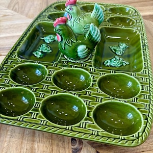 May include: A green ceramic egg tray with a woven basket design. The tray has egg-shaped and square compartments, and two decorative chicken figurines with red combs. The ceramic is glossy and reflects light.