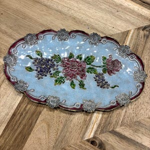 May include: An oval-shaped decorative tray featuring a light blue textured base with a central floral motif in shades of pink, purple, and green. The scalloped edge is outlined in deep red and accented with small, raised silver-toned flower details.