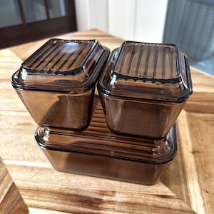 May include: Three rectangular storage containers made of translucent brown glass. Two smaller containers with ribbed lids sit atop one larger rectangular container. Each lid features a distinct linear pattern, and the glass has a smooth, tinted finish.