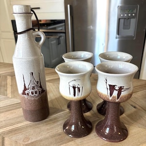 May include: A ceramic decanter with a cork stopper and four matching stemmed goblets. The set features a speckled cream upper section and a dark brown textured base. The decanter displays a brown cabin illustration, while the goblets show abstract brown designs.
