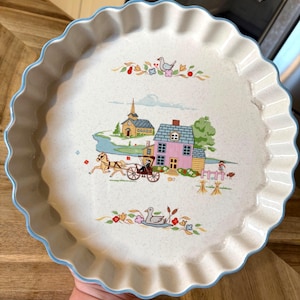 May include: A round, off-white ceramic quiche dish with a scalloped edge and a light blue rim. The centre features a colourful illustration of a pink house, a horse-drawn carriage, a church, and a pond, framed by floral and bird motifs.