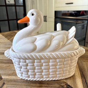 May include: A white ceramic lidded tureen shaped like a duck sitting in a woven basket. The duck has a bright orange beak and a black eye. The base features a textured basket-weave pattern with rope-style trim along the top and bottom edges.