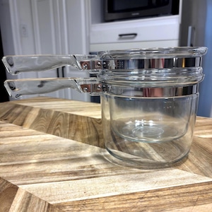 May include: A double boiler set consisting of two clear glass pots stacked vertically. Each pot features a transparent glass handle attached with a silver-coloured metal band. The glass is smooth and reflective, showing a clear view through the vessels.