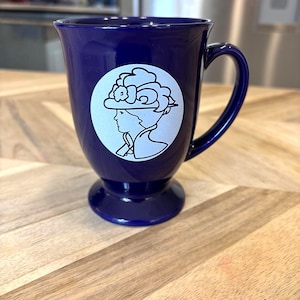 May include: A deep blue ceramic mug with a flared rim and a pedestal base. The front of the mug features a white circular decal depicting the profile of a person wearing a wide-brimmed hat with floral details, rendered in a simple line art style.