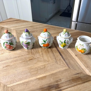 Peut inclure: Un ensemble de cinq pots en céramique blanche avec couvercles, chacun décoré d'illustrations de fruits colorés, notamment des pêches, du raisin, des oranges et des citrons. Les couvercles ont des poignées en forme de fruits.