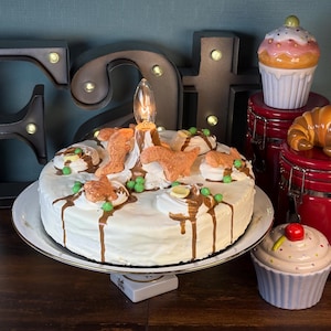 以下が含まれることがあります： チョコレートのドリップとオレンジ色の装飾が施された白いフロストケーキが、白いケーキスタンドに置かれています。装飾的なカップケーキとクロワッサンの瓶がケーキの隣にあります。「F」と「S」の文字が背景で点灯しています。
