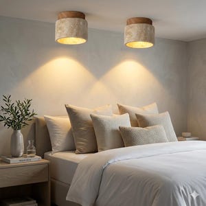 May include: Two cylindrical ceiling lights with a wood top and a textured, beige body are mounted on the ceiling. The lights illuminate a bed with white bedding and several beige pillows. A nightstand with a vase and a book is on the left.