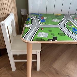 May include: A children's table and chair set featuring a light wood finish and white accents. The tabletop is covered in a green play mat graphic depicting a road network, trees, and buildings with the text PPP printed on a gray square.