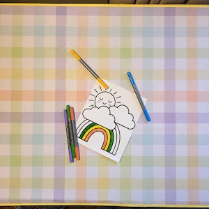 May include: A rectangular table with a pastel rainbow plaid pattern and yellow trim. On the surface sits a white paper with a black line drawing of a smiling sun, clouds, and a rainbow, accompanied by five colorful felt-tip markers.