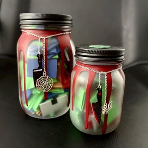May include: Two glass jars with black lids, featuring red paint drips down the sides. Each jar has a silver chain necklace with a black knife charm and a silver spiderweb or bat charm. The jars are filled with folded colorful paper notes.