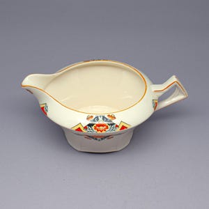 Puede incluir: Cremera de cerámica color crema con un patrón floral y geométrico decorativo en rojo, amarillo y azul. La cremera tiene un pico, un asa y un ribete dorado. Una pieza vintage para servir crema o salsas.
