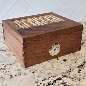 May include: A rectangular wooden humidor box featuring dark brown wood grain, visible finger joint corners, and a circular gold-toned analog hygrometer on the front. The lid includes a light-colored wooden grid insert with a geometric pattern.