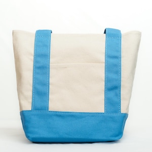 May include: A canvas tote bag with a cream-colored body and a bright blue base and straps. The straps are sewn with white stitching. The bag has a small pocket on the front.