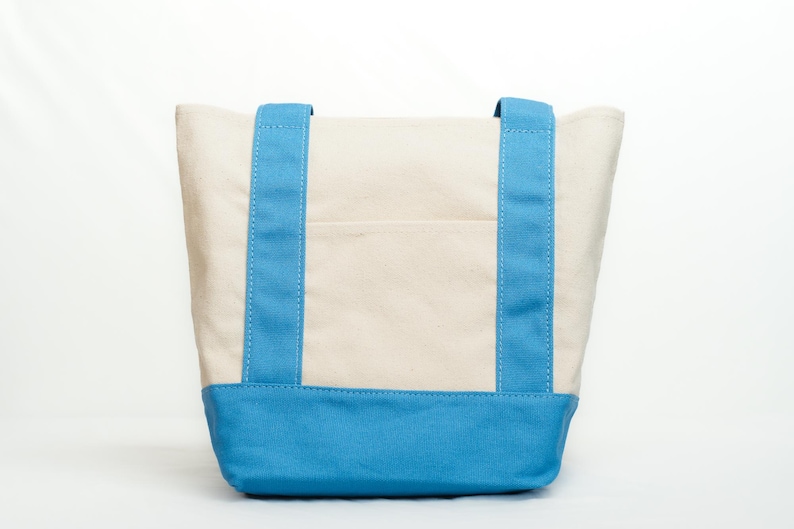 May include: A canvas tote bag with a cream-colored body and a bright blue base and straps. The straps are sewn with white stitching. The bag has a small pocket on the front.