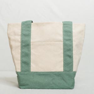 May include: A canvas tote bag with a cream-colored body and teal green accents. The bag features teal green handles and a teal green base. A pocket is visible on the front of the bag.
