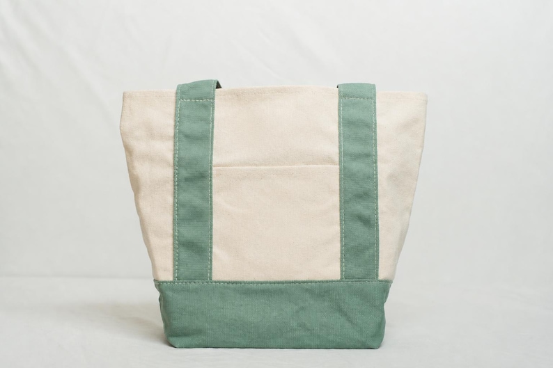 May include: A canvas tote bag with a cream-colored body and teal green accents. The bag features teal green handles and a teal green base. A pocket is visible on the front of the bag.
