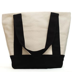 May include: A canvas tote bag with a cream-colored body and black accents. The bag features black handles and a black base. A small pocket is visible on the front of the bag.
