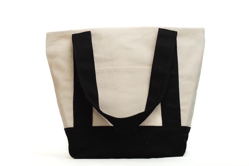 May include: A canvas tote bag with a cream-colored body and black accents. The bag features black handles and a black base. A small pocket is visible on the front of the bag.