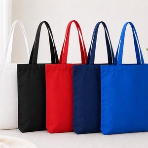 Op de afbeelding: Vijf canvas draagtassen op een rij, elk in een andere kleur. De kleuren zijn wit, zwart, rood, marineblauw en helderblauw. Elke tas heeft bijpassende handvatten.