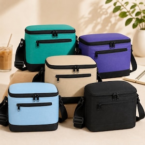 May include: Five insulated lunch bags in various colors: teal, purple, tan, light blue, and black. Each bag has a black base, a zippered front pocket, and a black shoulder strap. The bags are square-shaped and appear to be made of a durable fabric.