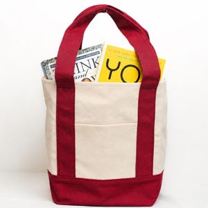 May include: A canvas tote bag with a cream-colored body and deep red accents on the base, handles, and side stripes. The bag contains two books, one with the title "Think and Grow Rich" and another with the word "YO" on the cover.