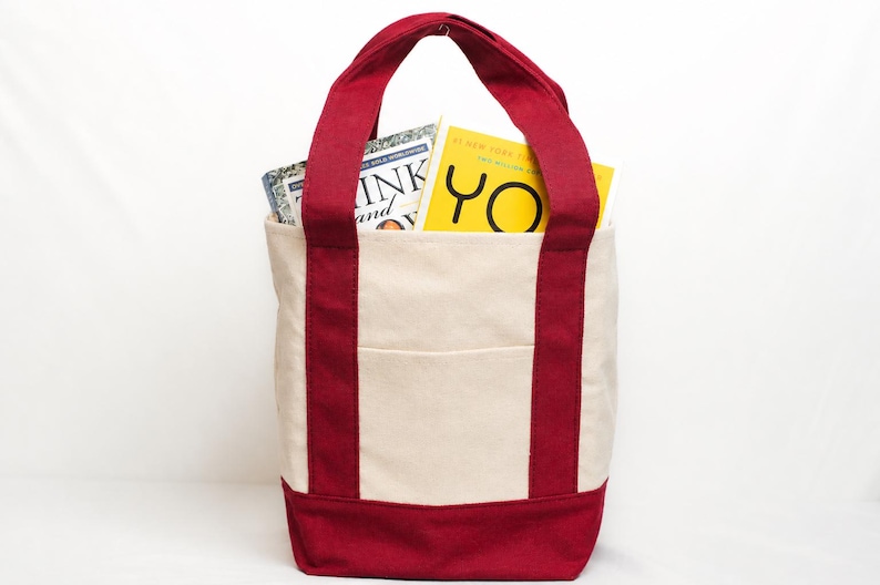 May include: A canvas tote bag with a cream-colored body and deep red accents on the base, handles, and side stripes. The bag contains two books, one with the title "Think and Grow Rich" and another with the word "YO" on the cover.