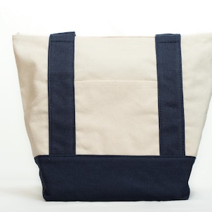 May include: Cream-colored canvas tote bag with navy blue accents. The bag features navy blue handles and a navy blue base. A small pocket is visible on the front of the bag. The bag is designed for carrying items.