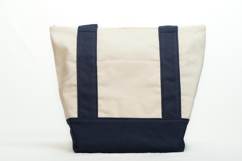 May include: Cream-colored canvas tote bag with navy blue accents. The bag features navy blue handles and a navy blue base. A small pocket is visible on the front of the bag. The bag is designed for carrying items.
