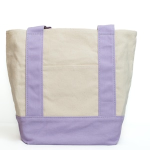 May include: A canvas tote bag with a light beige body and lavender accents. The bag features lavender handles and a lavender base. A small pocket is visible on the front of the bag. The bag is designed for carrying items.