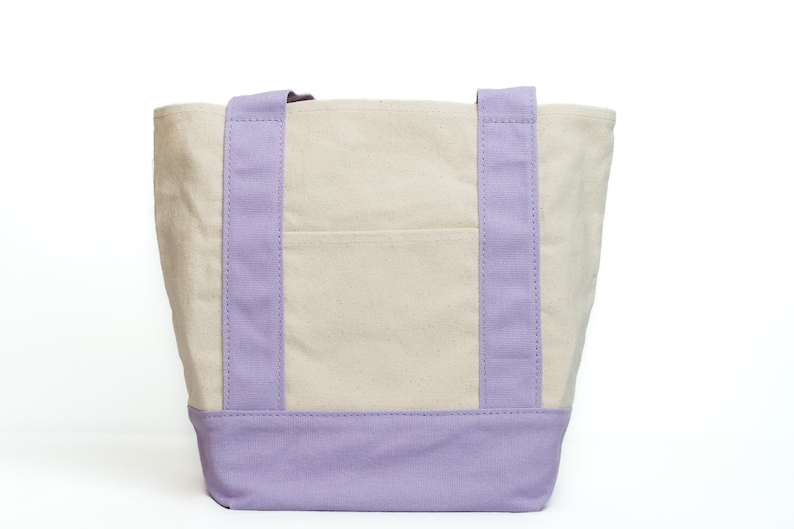 May include: A canvas tote bag with a light beige body and lavender accents. The bag features lavender handles and a lavender base. A small pocket is visible on the front of the bag. The bag is designed for carrying items.