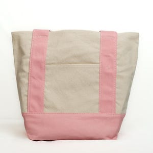 May include: A canvas tote bag with a light beige body, a pink base, and pink straps. The bag has a small pocket on the front. The tote bag is designed for carrying items.