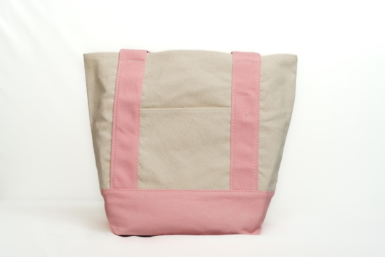 May include: A canvas tote bag with a light beige body, a pink base, and pink straps. The bag has a small pocket on the front. The tote bag is designed for carrying items.