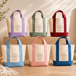 May include: Six small canvas tote bags arranged in two rows. Each bag features a cream-colored body with a colored base and handles in lavender, sage green, red, light blue, pink, or dark blue. Each bag has a front pocket with the text YOUR DESIGN HERE.