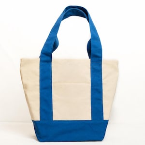 May include: A canvas tote bag with a cream-colored body and vibrant blue accents. The bag features blue handles and a blue base, with vertical blue stripes on the front and back. The bag is designed with a simple, functional style.