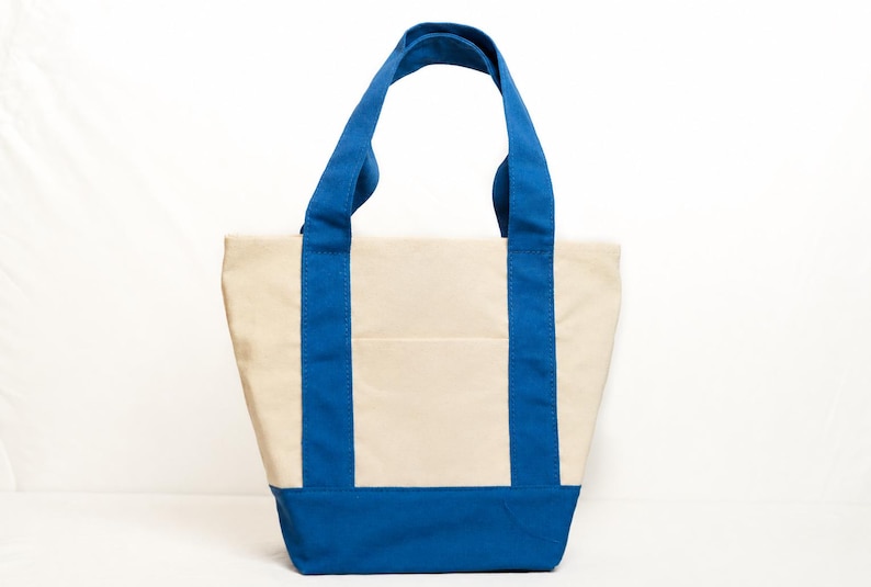May include: A canvas tote bag with a cream-colored body and vibrant blue accents. The bag features blue handles and a blue base, with vertical blue stripes on the front and back. The bag is designed with a simple, functional style.