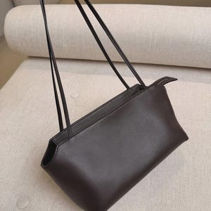May include: A dark brown shoulder bag with a smooth, solid-colored exterior, a rectangular base, and two thin, long straps attached to the top. The bag features a zippered closure along the top edge and clean, structured stitching along the seams.