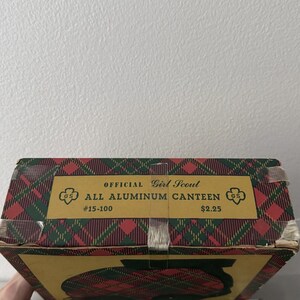 Puede incluir: Caja de cantimplora vintage de las Girl Scouts con un patr&oacute;n de cuadros rojos, verdes y amarillos. La caja presenta el texto "OFFICIAL Girl Scout ALL ALUMINUM CANTEEN" y el logotipo de las Girl Scouts.