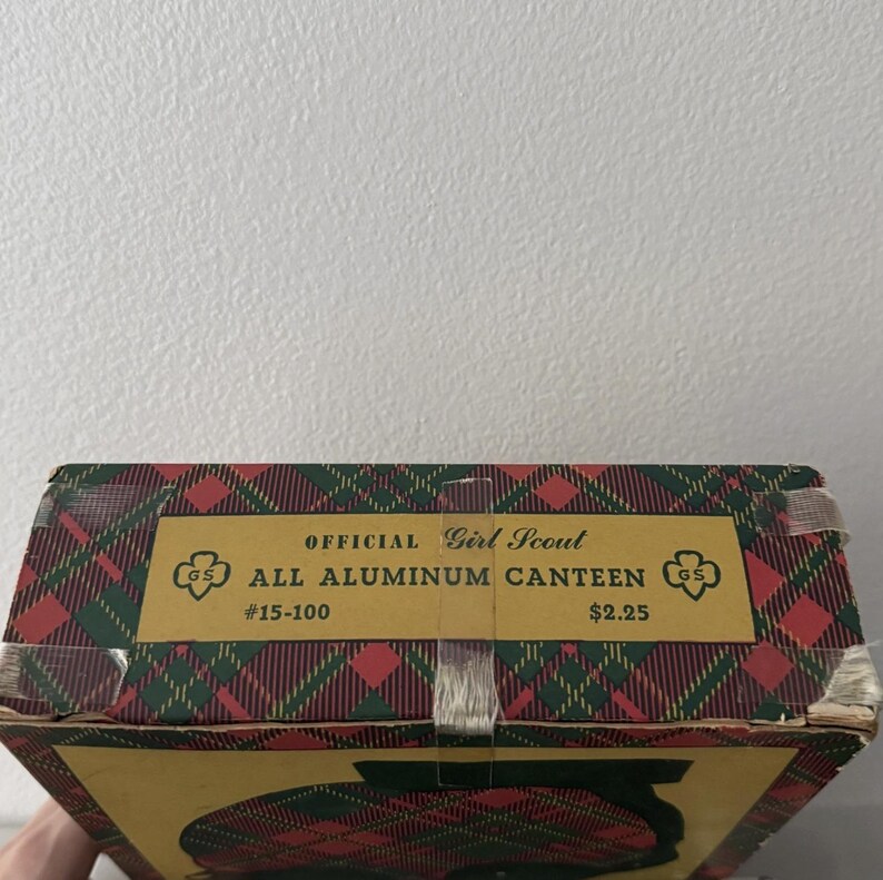 Puede incluir: Caja de cantimplora vintage de las Girl Scouts con un patr&oacute;n de cuadros rojos, verdes y amarillos. La caja presenta el texto "OFFICIAL Girl Scout ALL ALUMINUM CANTEEN" y el logotipo de las Girl Scouts.