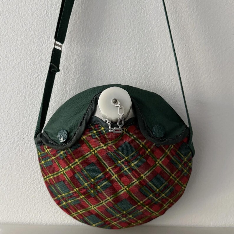 Puede incluir: Cantimplora vintage con un estampado de cuadros rojos, verdes y amarillos en la mitad inferior. La parte superior es de color verde oscuro y tiene una tapa blanca sujeta con una cadena. Se adjunta una correa verde oscuro para llevarla.