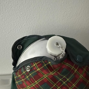 Puede incluir: Una cantimplora vintage con cuerpo de metal y tap&oacute;n blanco sujeto por una cadena. La cantimplora est&aacute; alojada en una funda de tela a cuadros verdes y rojos con cierres de bot&oacute;n a presi&oacute;n. La funda tiene una solapa y ribete verdes.