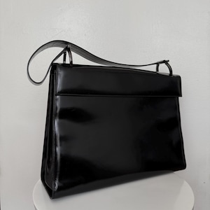 May include: A structured black handbag with a glossy, patent-like finish. The bag features a rectangular silhouette, a wide top flap, and a single arched handle attached with metal hardware. The surface is smooth and reflective with clean edges.