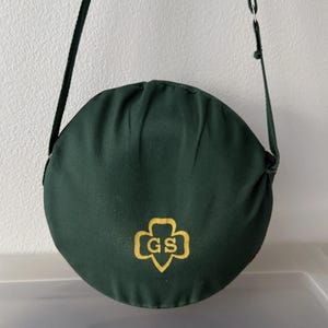 Puede incluir: Un bolso de hombro circular verde oscuro con una correa larga negra. El bolso presenta un emblema dorado con las letras "GS" dentro de una forma de tr&eacute;bol. El bolso est&aacute; hecho de un material suave, posiblemente similar a la lona.