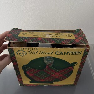 Puede incluir: Cantimplora vintage de las Girl Scouts en su caja original. La caja presenta un estampado de cuadros rojos, verdes y dorados. Las palabras "Official Girl Scout Canteen" est&aacute;n impresas en la caja. La cantimplora es visible dentro.