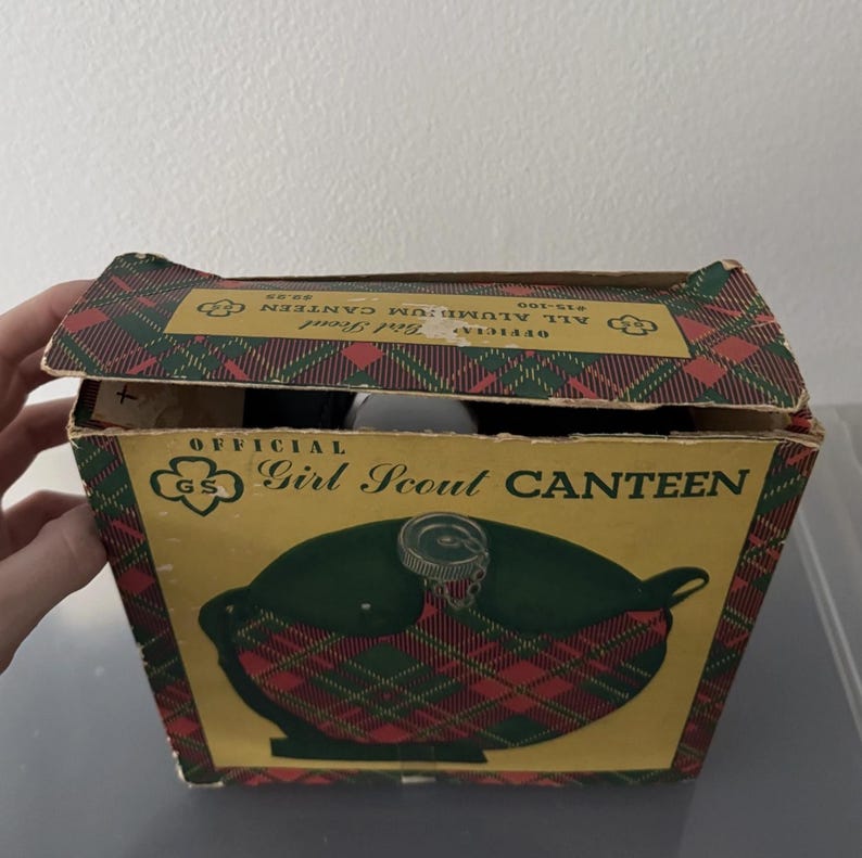 Puede incluir: Cantimplora vintage de las Girl Scouts en su caja original. La caja presenta un estampado de cuadros rojos, verdes y dorados. Las palabras "Official Girl Scout Canteen" est&aacute;n impresas en la caja. La cantimplora es visible dentro.