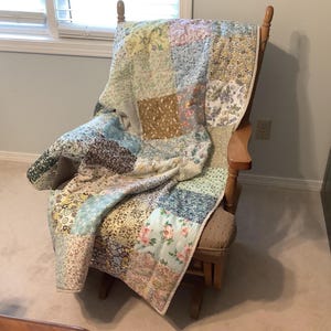 May include: A patchwork quilt featuring a variety of floral and geometric fabric squares in shades of blue, yellow, pink, and brown. The quilt is draped over a wooden rocking chair with a padded seat and backrest, showcasing a quilted texture.