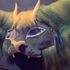 Może przedstawiać: Niebiesko-zielona maska fantasy z białymi rogami i kremową grzywą. Maska posiada spiralny wz&oacute;r na lewym oku i siatkową osłonę oka. Maska ma koci nos i kły.