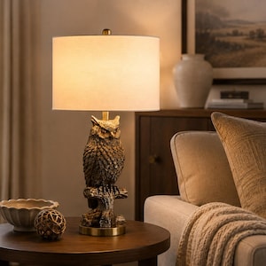 May include: A table lamp featuring a base sculpted as a detailed brown owl perched on a tree branch. The lamp has a cylindrical off-white fabric shade that emits a warm glow, supported by a brass-toned metal stem and a circular base.