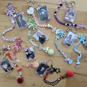 Könnte beinhalten: Eine Sammlung von acht rechteckigen Acryl-Fotokeychains mit Porträts. Jeder Schlüsselanhänger ist an einer Perlenkette mit verschiedenen Anhängern befestigt, darunter Sterne, Schleifen, Pompons und Buchstabenperlen, die Namen wie YOONGI, J-HOPE und TAEHYUNG bilden.