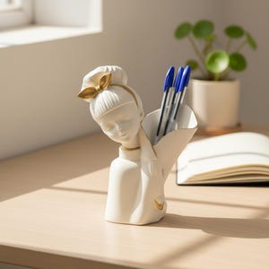 Puede incluir: Un organizador de escritorio de cerámica blanca con forma de busto estilizado con un lazo de color dorado en el cabello. La figura cuenta con una sección hueca en la parte posterior diseñada para sostener bolígrafos, lápices u otros artículos.